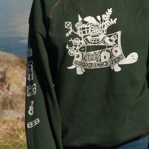 Könnte beinhalten: Ein dunkelgrüner Pullover mit einem weißen Aufdruck einer Schildkröte, die ein Haus auf ihrem Rücken trägt. Die Schildkröte ist von verschiedenen anderen kleinen weißen Designs umgeben.