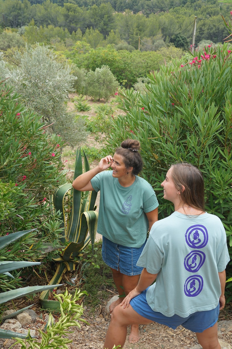 May include: Two people wearing light blue t-shirts with purple designs. The person on the left is wearing a denim skirt and the person on the right is wearing denim shorts. They are standing in a garden with green plants and flowers.