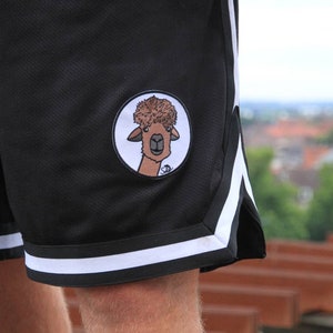 May include: Black athletic shorts with white stripes and a circular patch featuring a cartoon llama with a fluffy brown hairstyle. The shorts have a mesh texture and are designed for sports or casual wear.