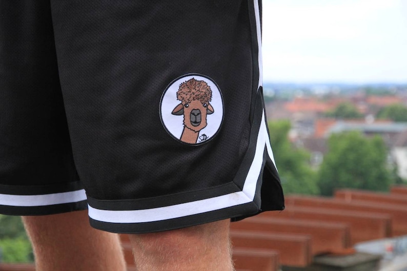 May include: Black athletic shorts with white stripes and a circular patch featuring a cartoon llama with a fluffy brown hairstyle. The shorts have a mesh texture and are designed for sports or casual wear.