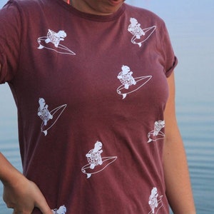 May include: A maroon t-shirt with a white graphic print of turtles surfing. The turtles are wearing sunglasses and have a happy expression.