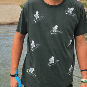 Puede incluir: Una camiseta verde oscuro con un estampado gráfico blanco de tortugas montando tablas de surf. La camiseta tiene cuello redondo y mangas cortas.