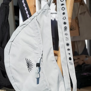 May include: A light gray sling bag with a black sunburst graphic and a strap adorned with various sun and moon illustrations. The bag is hanging on a wooden rack, showcasing its design and features.