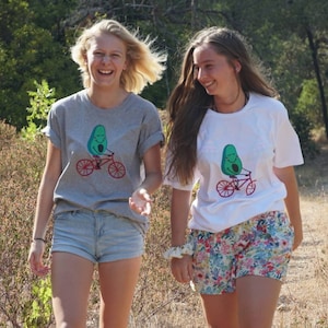 Pode incluir: Duas pessoas vestindo camisetas cinza e branca com um desenho de um abacate de desenho animado andando de bicicleta. A pessoa que est&aacute; vestindo a camiseta cinza est&aacute; sorrindo e olhando para a c&acirc;mera. A pessoa que est&aacute; vestindo a camiseta branca est&aacute; olhando para a pessoa que est&aacute; vestindo a camiseta cinza. Ambas as pessoas est&atilde;o vestindo shorts.