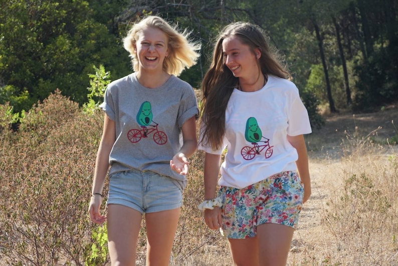 Pode incluir: Duas pessoas vestindo camisetas cinza e branca com um desenho de um abacate de desenho animado andando de bicicleta. A pessoa que est&aacute; vestindo a camiseta cinza est&aacute; sorrindo e olhando para a c&acirc;mera. A pessoa que est&aacute; vestindo a camiseta branca est&aacute; olhando para a pessoa que est&aacute; vestindo a camiseta cinza. Ambas as pessoas est&atilde;o vestindo shorts.