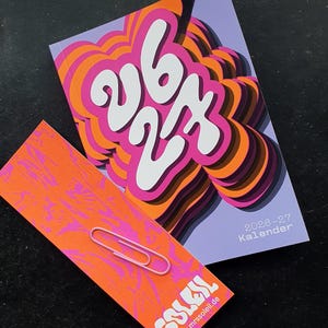 May include: A calendar and a card with a pink paperclip. The calendar has a purple background with the numbers "26 27" in white, and the word "Kalender" in the corner. The card is orange with pink swirls and the word "SOLEIL" in white.
