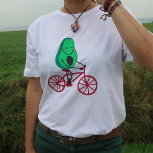 Pode incluir: Camiseta branca com um personagem de abacate verde a andar de bicicleta vermelha. O abacate tem uma cara sorridente e usa um capacete vermelho.