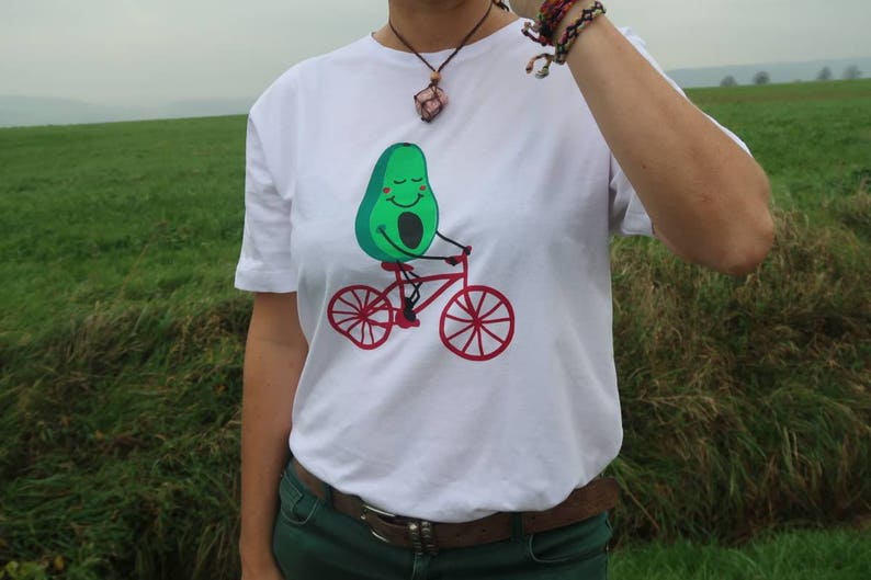 Pode incluir: Camiseta branca com um personagem de abacate verde a andar de bicicleta vermelha. O abacate tem uma cara sorridente e usa um capacete vermelho.