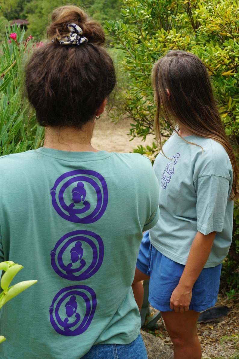 May include: Two people wearing light blue t-shirts with purple graphic designs on the back. The person on the left has a bun in their hair and is wearing blue jeans. The person on the right is wearing blue shorts.