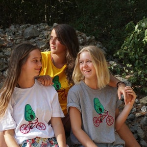 Pode incluir: Tr&ecirc;s amigas est&atilde;o vestindo camisetas com um desenho animado de um abacate andando de bicicleta. O abacate tem um rosto sorridente. As amigas est&atilde;o sentadas em uma pilha de pedras em um ambiente de floresta.