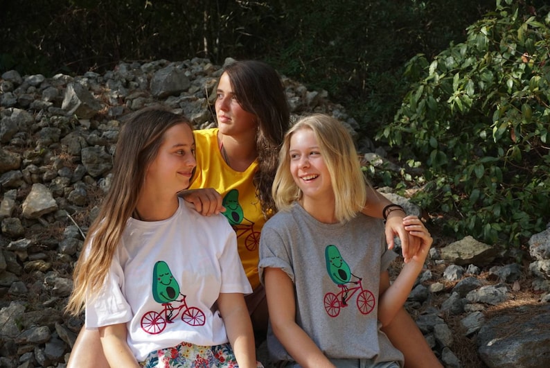 Pode incluir: Tr&ecirc;s amigas est&atilde;o vestindo camisetas com um desenho animado de um abacate andando de bicicleta. O abacate tem um rosto sorridente. As amigas est&atilde;o sentadas em uma pilha de pedras em um ambiente de floresta.