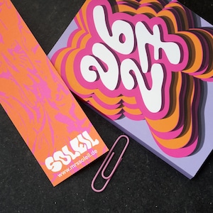 May include: An orange bookmark with pink patterns and the white word "SOLEIL". A calendar with the text "627" in a layered design, and the text "2028-27 Kalender". A pink paperclip is next to the calendar and bookmark.