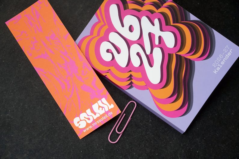 May include: An orange bookmark with pink patterns and the white word "SOLEIL". A calendar with the text "627" in a layered design, and the text "2028-27 Kalender". A pink paperclip is next to the calendar and bookmark.