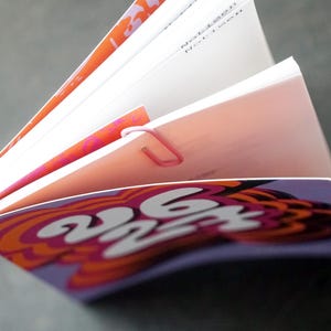 May include: An open book with pages fanned out, held together by a pink paper clip. The cover features a purple and orange psychedelic design with the numbers "262". The pages have orange and pink accents.