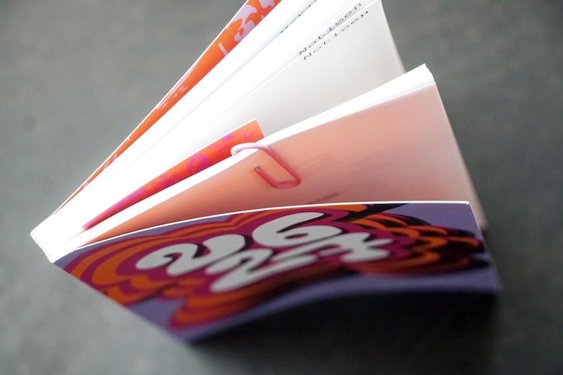 May include: An open book with pages fanned out, held together by a pink paper clip. The cover features a purple and orange psychedelic design with the numbers "262". The pages have orange and pink accents.
