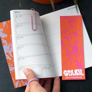 May include: An open notebook with a pink paperclip, a bookmark, and a patterned cover. The bookmark and cover are orange with a pink design. The bookmark has the word "SOLEIL" and the website "www.mrssoleil.de".