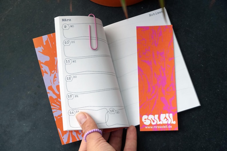 May include: An open notebook with a pink paperclip, a bookmark, and a patterned cover. The bookmark and cover are orange with a pink design. The bookmark has the word "SOLEIL" and the website "www.mrssoleil.de".