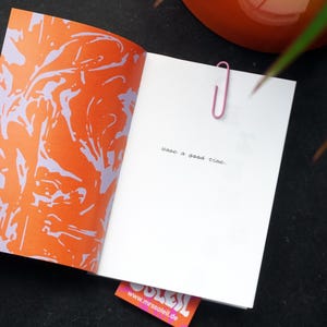 May include: An open book with an orange and purple abstract design on the left page. The right page is white with the handwritten text "have a good time." A pink paperclip holds the pages together. A small pink and orange tag is visible.