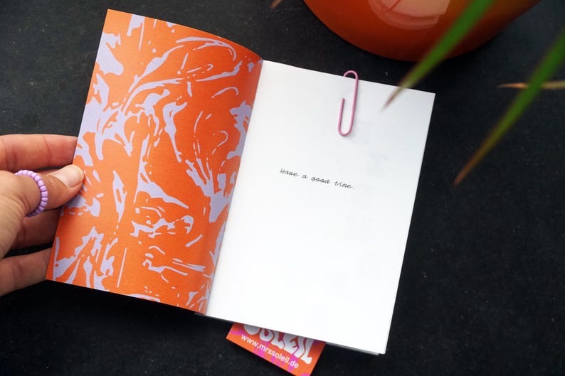 May include: An open book with an orange and purple abstract design on the left page. The right page is white with the handwritten text "have a good time." A pink paperclip holds the pages together. A small pink and orange tag is visible.