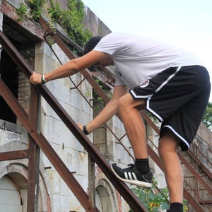 May include: A person climbing a rusty metal structure attached to a weathered stone building. The person is wearing a white t-shirt, black shorts with white trim, and black sneakers. The building has arched openings and a partially collapsed roof.