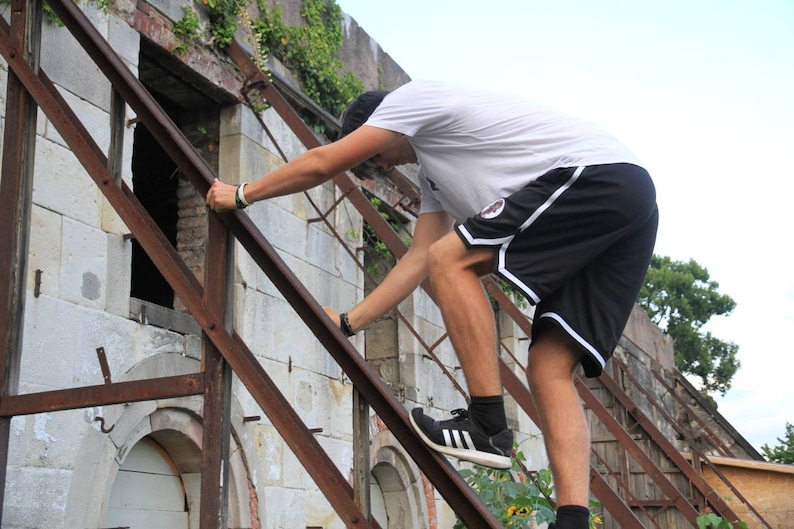 May include: A person climbing a rusty metal structure attached to a weathered stone building. The person is wearing a white t-shirt, black shorts with white trim, and black sneakers. The building has arched openings and a partially collapsed roof.