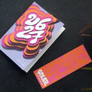 May include: A calendar with a layered design in pink, orange, and white, displaying the numbers "26 27". A matching bookmark with the word "SOLEIL" and website address is also present. The calendar is labelled "2026-27 Kalender".