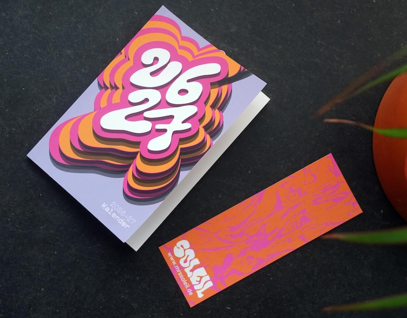 May include: A calendar with a layered design in pink, orange, and white, displaying the numbers "26 27". A matching bookmark with the word "SOLEIL" and website address is also present. The calendar is labelled "2026-27 Kalender".