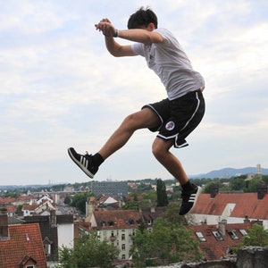 May include: A person in mid-air, performing a jump. They are wearing a white t-shirt, black shorts with white trim, and black sneakers with white stripes. The background shows a cityscape under a cloudy sky.
