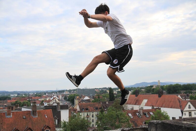 May include: A person in mid-air, performing a jump. They are wearing a white t-shirt, black shorts with white trim, and black sneakers with white stripes. The background shows a cityscape under a cloudy sky.