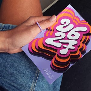 May include: A vibrant calendar featuring the numbers "26 27" in white, set against a layered design of orange, pink, and purple. The calendar is held in a hand wearing a beaded ring, with denim jeans in the background.