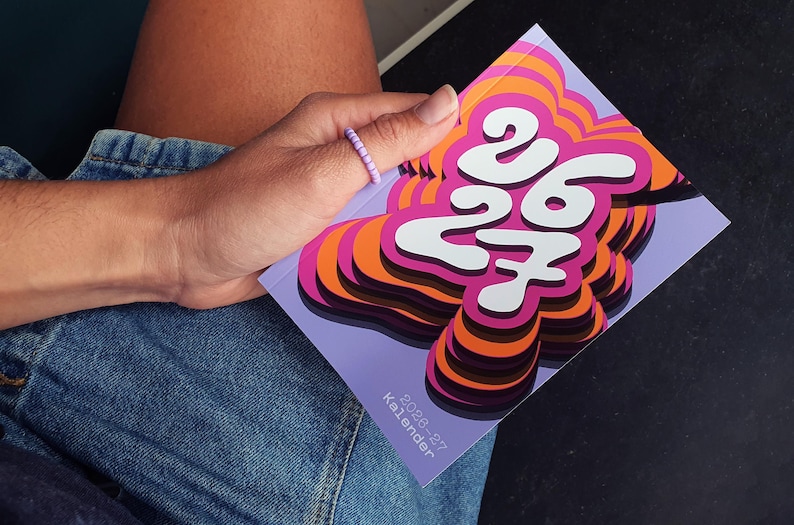 May include: A vibrant calendar featuring the numbers "26 27" in white, set against a layered design of orange, pink, and purple. The calendar is held in a hand wearing a beaded ring, with denim jeans in the background.