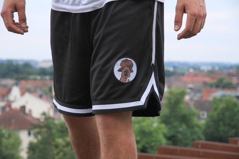 May include: Black athletic shorts with white stripes along the sides and bottom hem. A circular patch featuring a cartoon llama is on the left leg. The shorts are made of a mesh material.