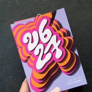 May include: A vibrant calendar with a layered design. The numbers "26 27" are in white against a pink, orange, and purple background. The text "2026-27 Kalender" is visible. The calendar is held in a hand.