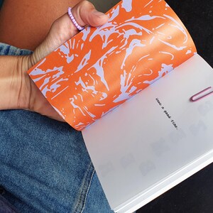 May include: Open notebook with an orange and purple abstract design on the inside cover. The page reads "have a good time," held open with a pink paper clip. The person is wearing a denim skirt.
