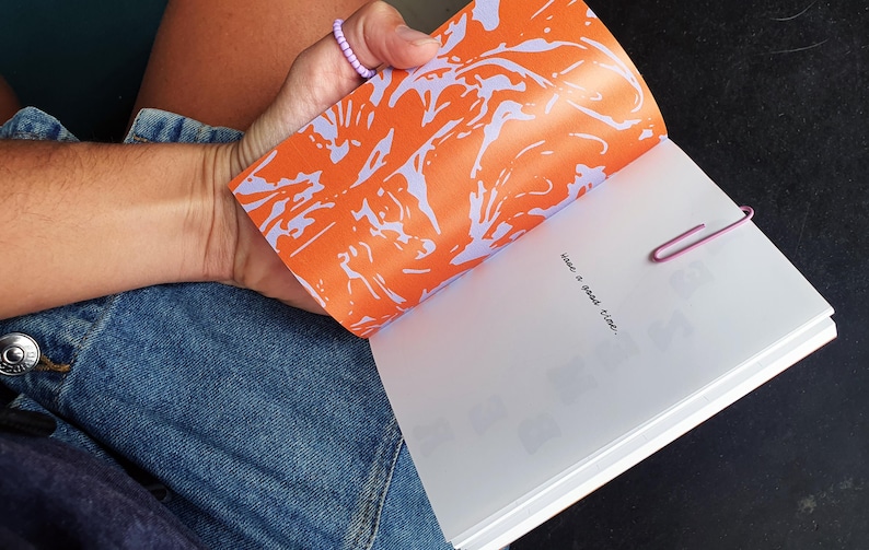 May include: Open notebook with an orange and purple abstract design on the inside cover. The page reads "have a good time," held open with a pink paper clip. The person is wearing a denim skirt.