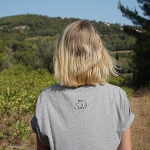 Pode incluir: Uma mulher vestindo uma camiseta cinza com o texto "Mrs. Smith" nas costas. Ela est&aacute; em p&eacute; em um campo com vista para uma floresta ao fundo.