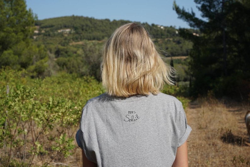Pode incluir: Uma mulher vestindo uma camiseta cinza com o texto "Mrs. Smith" nas costas. Ela est&aacute; em p&eacute; em um campo com vista para uma floresta ao fundo.