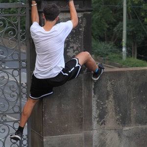 May include: A person wearing a white t-shirt and black shorts with white trim is climbing a concrete wall. The person is wearing black sneakers with white soles and blue socks. A decorative metal gate is in the background.