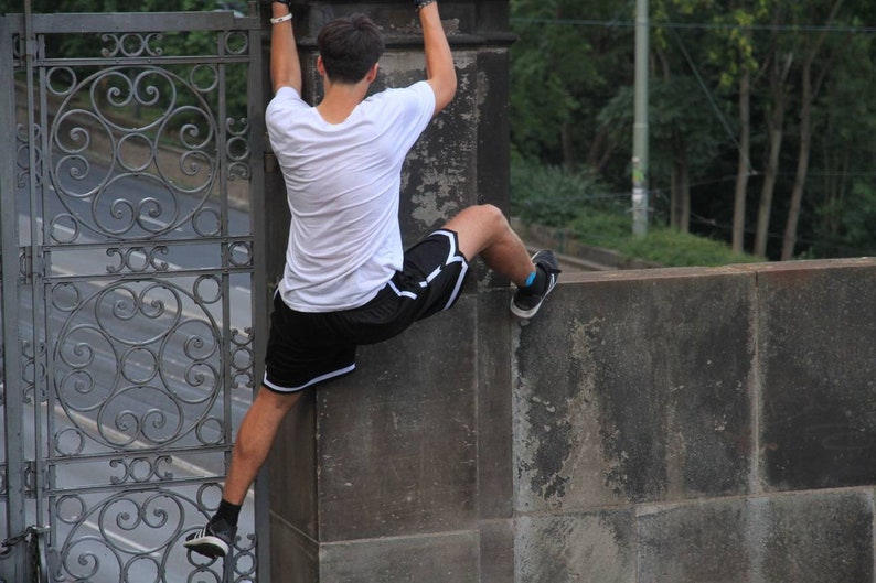 May include: A person wearing a white t-shirt and black shorts with white trim is climbing a concrete wall. The person is wearing black sneakers with white soles and blue socks. A decorative metal gate is in the background.