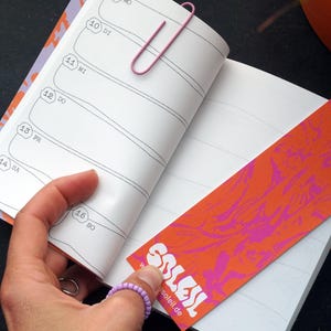 May include: An open notebook with lined pages and a pink paperclip. A bookmark with an orange and pink abstract design is inserted. The word "SOLEIL" is visible on the bookmark, along with the website soleil.de.