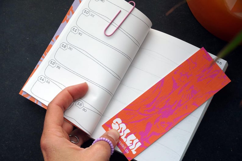 May include: An open notebook with lined pages and a pink paperclip. A bookmark with an orange and pink abstract design is inserted. The word "SOLEIL" is visible on the bookmark, along with the website soleil.de.