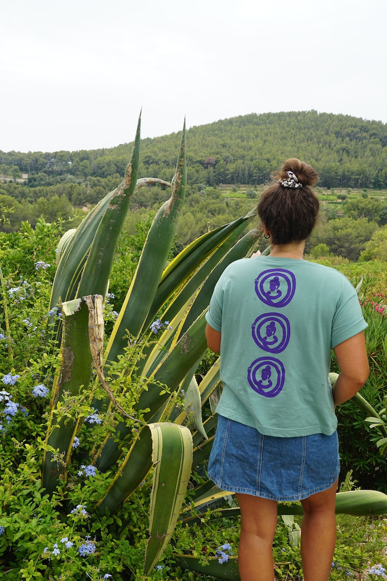 May include: A woman wearing a teal t-shirt with three purple circles on the back. Each circle has a stylized image of a person with their hair in a bun. The woman is wearing a denim skirt and is standing in front of a large agave plant.