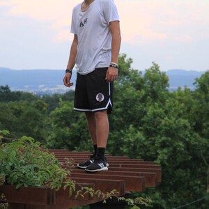 May include: Black athletic shorts with white trim and a circular emblem. The shorts are paired with a white t-shirt and black sneakers. The image is taken outdoors with a background of green trees and a cloudy sky.