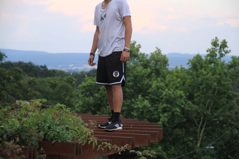 May include: Black athletic shorts with white trim and a circular emblem. The shorts are paired with a white t-shirt and black sneakers. The image is taken outdoors with a background of green trees and a cloudy sky.
