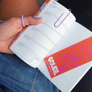 May include: An open planner with a pink paperclip and a bookmark. The bookmark has the text "SOLES" and a vibrant design. The planner's page shows dates and lines. The planner is held in a person's hand.