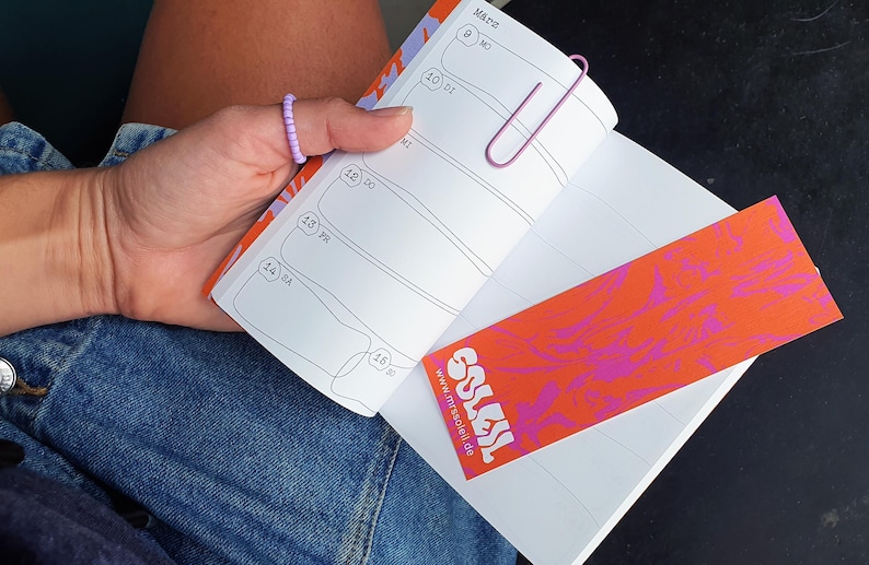 May include: An open planner with a pink paperclip and a bookmark. The bookmark has the text "SOLES" and a vibrant design. The planner's page shows dates and lines. The planner is held in a person's hand.