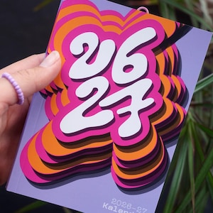 May include: A calendar with a lilac cover displaying the numbers "26 27" in white, with a layered pink and orange design. The text "2026-27 Kalender" is at the bottom. The calendar is held in a hand.