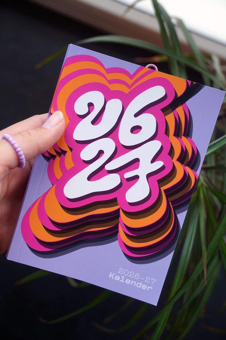 May include: A calendar with a lilac cover displaying the numbers "26 27" in white, with a layered pink and orange design. The text "2026-27 Kalender" is at the bottom. The calendar is held in a hand.