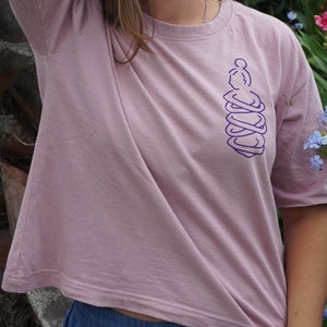 May include: A light pink t-shirt with a purple graphic of a person hugging themself. The shirt has short sleeves and a relaxed fit.