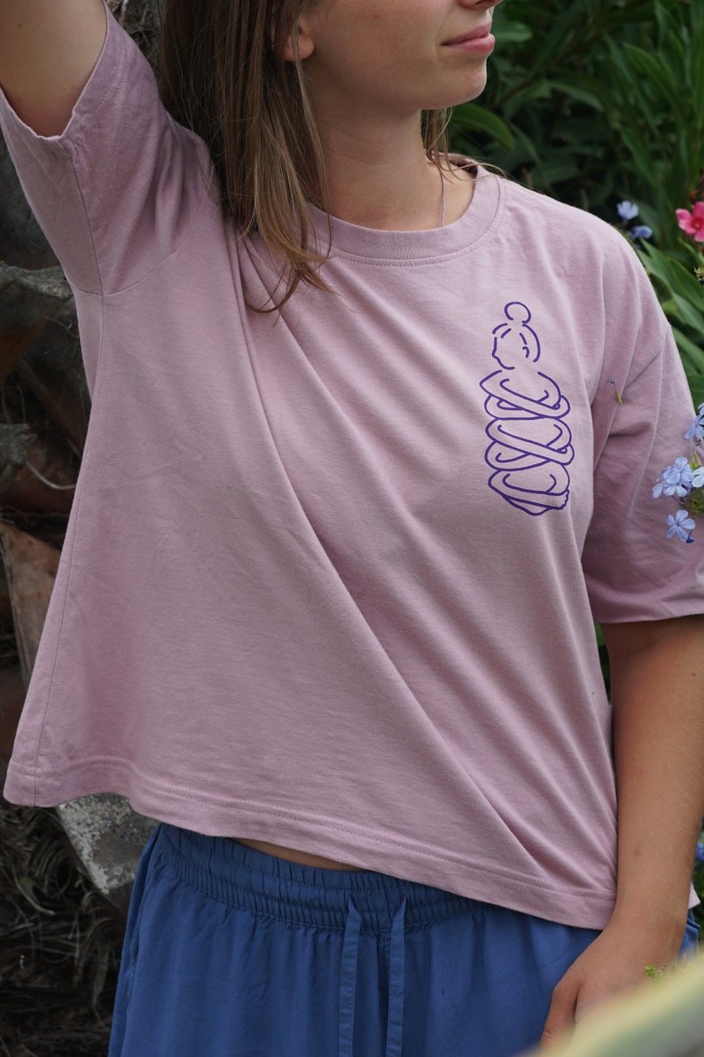 May include: A light pink t-shirt with a purple graphic of a person hugging themself. The shirt has short sleeves and a relaxed fit.
